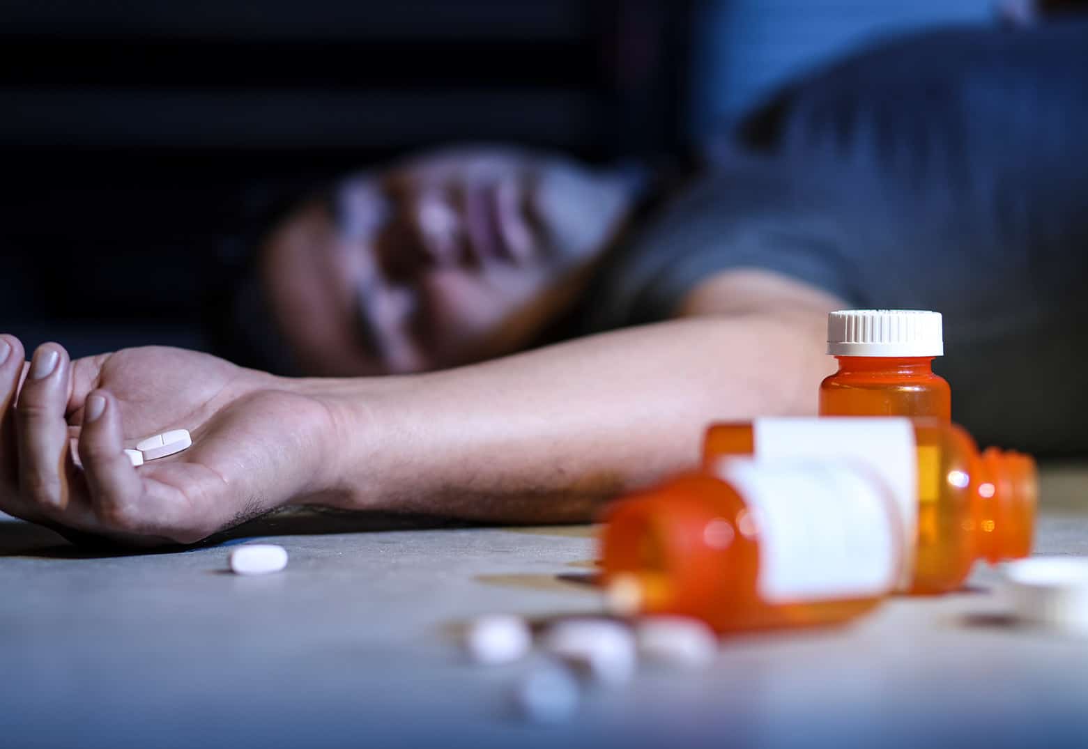 A male who is dead from an overdose with pills in his hands and empty pill bottles around him.
