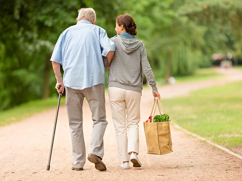 A female family caregiver walking beside an older male with a cane.