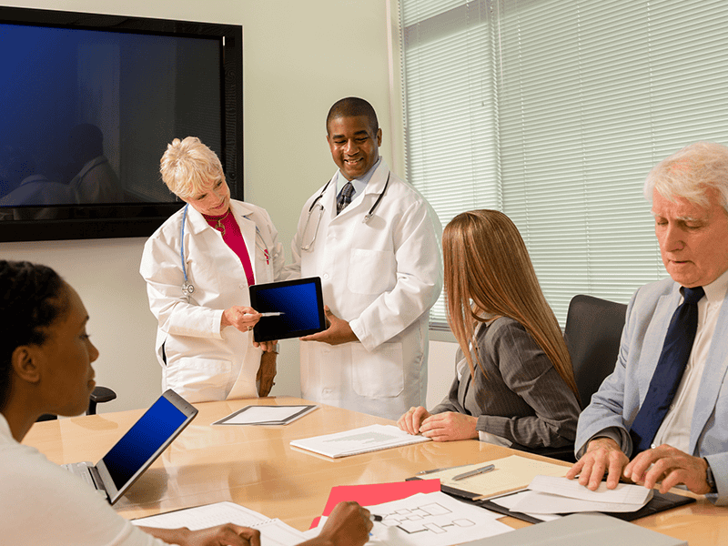 Physician-led care team and executives reviewing data on a tablet during a strategy meeting about value-based care challenges.
